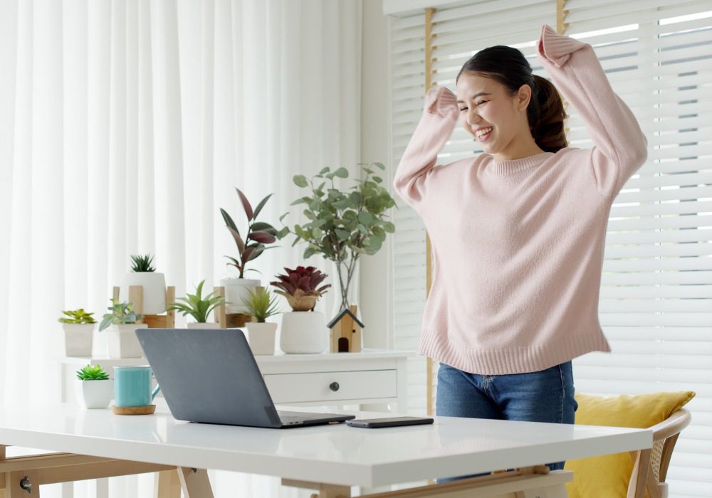 A female writer in her study wearing a pink jumper, looking at her laptop while standing up and cheering in celebration.